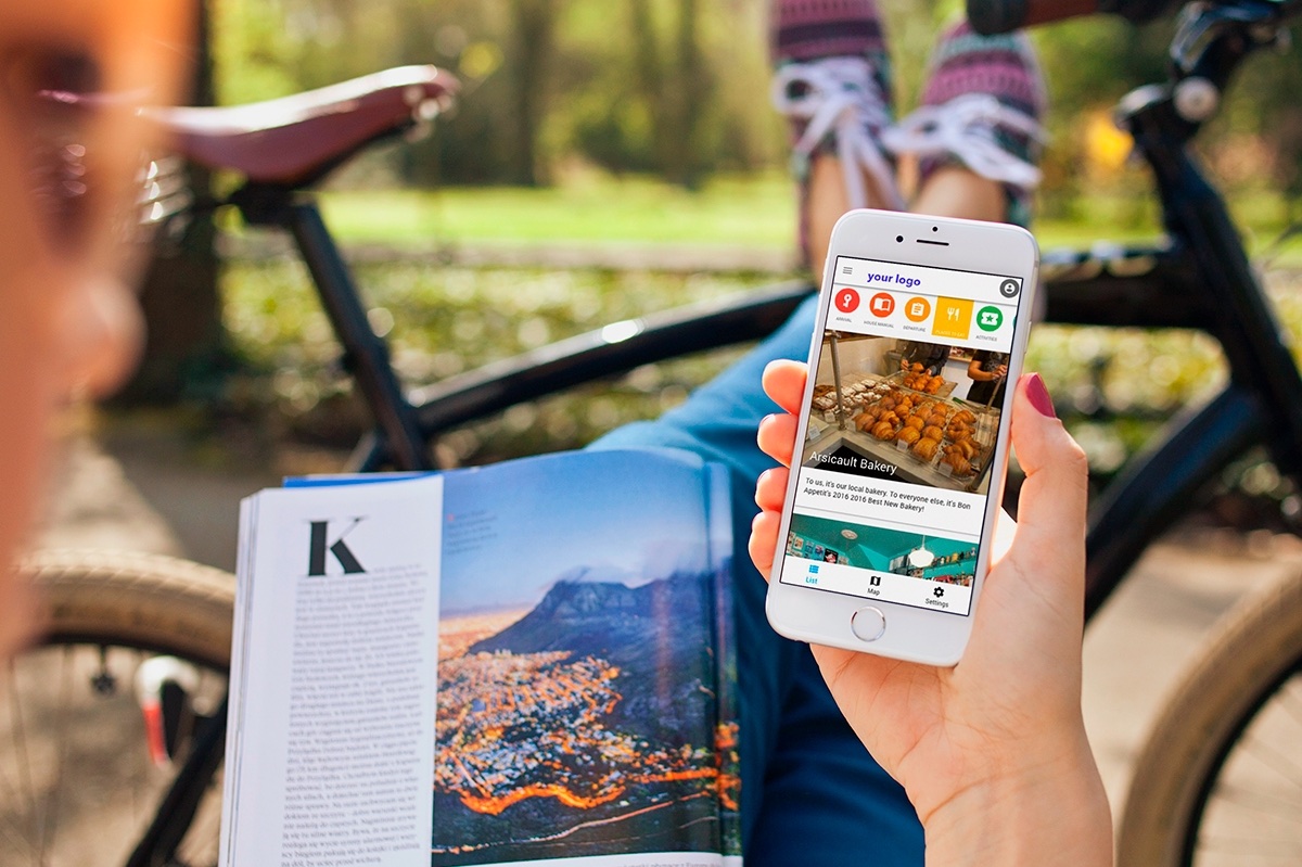 A person looking at their Hostfully digital guidebook on their phone while sitting outside in a park and reading a magazine