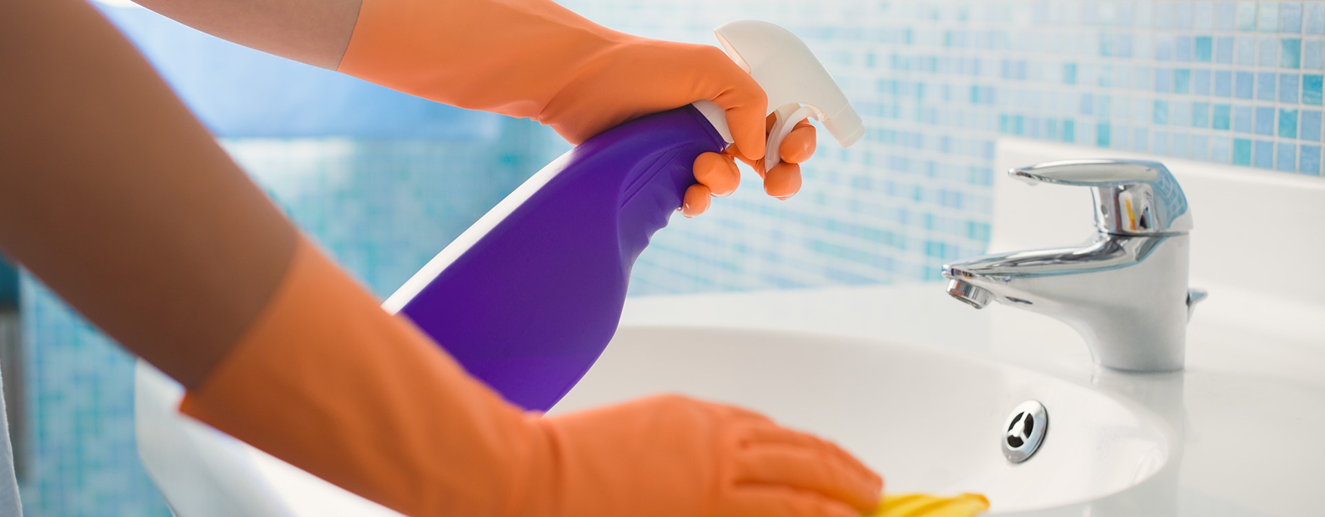 A person wearing orange gloves spraying down and cleaning a bathroom sink